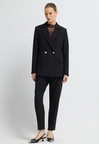 Czarna dwurzędowa marynarka z złotymi guzikami, dopasowane spodnie w kant oraz czarne czółenka na obcasie z wydłużonym noskiem. Gładki materiał i strukturalny design.