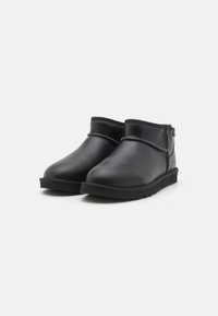 Botas de cuero negro hasta el tobillo con punteras redondeadas, acento de costura alrededor de la parte superior y suelas de goma planas. Diseño y textura minimalista.