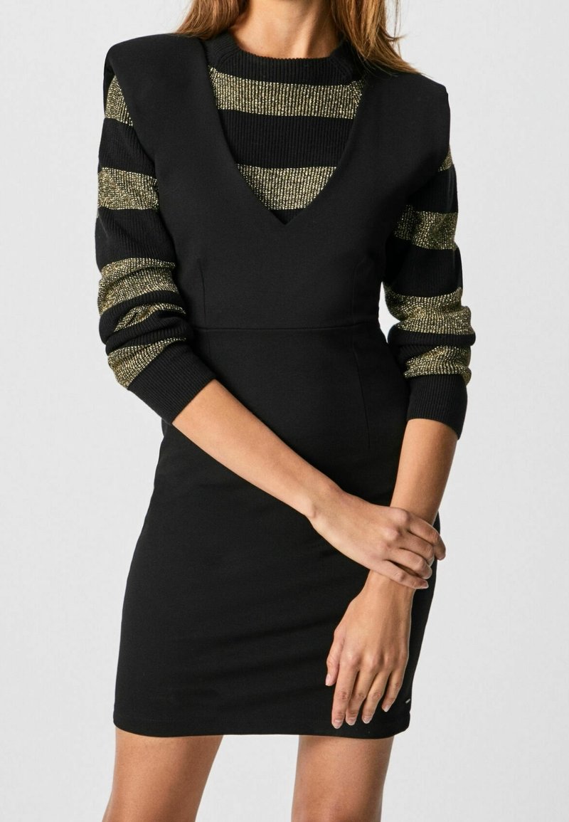 Femme portant une robe noire sans manches par-dessus un pull à rayures noires et dorées à manches longues, les bras croisés devant.