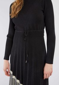 Schwarzes geripptes Strickkleid mit V-Ausschnitt, figurbetontem Oberteil, tailliertem Bund und plissiertem Rock. Mit einem Hahnentrittmuster am Saum.