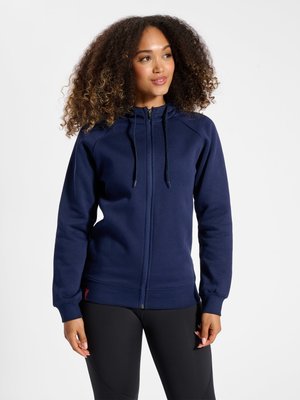 Kobieta z kręconymi włosami, ubrana w granatową bluzę rozpinaną na zamek i czarne legginsy, stojąca na jednolitym białym tle.