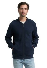 Granatowa kurtka na zamek z teksturowaną powierzchnią, ściągaczami na mankietach i dwoma bocznymi kieszeniami. Posiada okrągły kołnierz oraz subtelne logo.