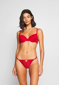 Rdeča čipkasta spodnje perilo komplet z nedrčkom z oblazinjenjem in nazobčanimi robovi ter ujemajočimi se tangicami. Vidna sta prosojna tekstura in detajli z cvetličnim vzorcem.