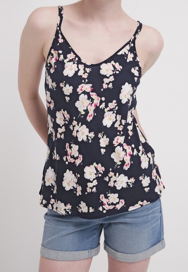 Personne portant un haut sans manches noir à motifs floraux et un short en jean retroussé, debout avec les bras derrière le dos sur un fond uni.