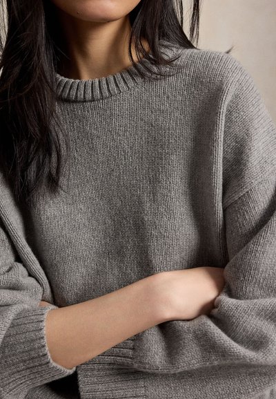 Γυναίκα με μακριά σκούρα μαλλιά που φορά γκρι πλεκτό πουλόβερ, με σταυρωμένα τα χέρια, εμφανίζεται από τους ώμους έως τη μέση σε ουδέτερο φόντο.