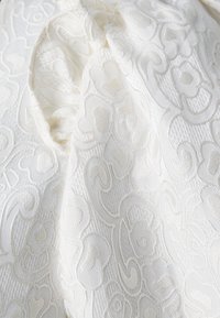 Tessuto bianco con intricati motivi floreali e swirl in jacquard in rilievo che creano pieghe di superficie decorative e testurizzate.