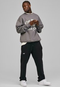 Grå hettegenser med hvit grafisk tekst, svarte joggebukser og hvite sneakers. Avslappet passform, bomullsmateriale, ribbede mansjetter og snøring i livet.