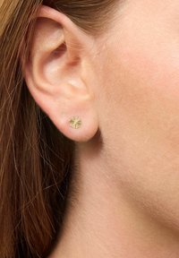 Goldene Ohrstecker mit floralen Motiven und strukturierten Blütenblättern, vor einem hellen Hautton und braunen Haaren. Eine Nahaufnahme betont das filigrane Design.