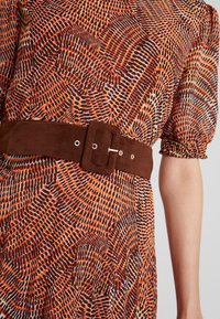 Langärmelig jurk met een oranje en bruine geometrische print, voorzien van een bruine suedeband met een rechthoekige gesp en zilverkleurige hardware.