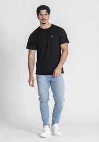 Camiseta negra de manga corta con un pequeño logotipo, combinada con jeans de color azul claro y zapatillas blancas sobre un fondo neutro.