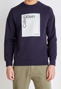 Marinblå sweatshirt med rund hals, långa ärmar och ribbade muddars. Har en vit rektangulär grafik med "Calvin Klein" och "Est. 1968."