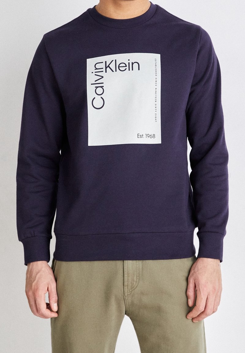 Marinblå sweatshirt med rund hals, långa ärmar och ribbade muddars. Har en vit rektangulär grafik med "Calvin Klein" och "Est. 1968."