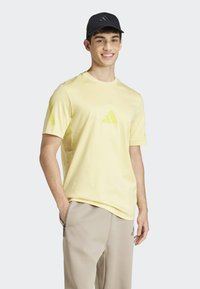 Žluté tričko s krátkým rukávem a černým trojúhelníkovým logem na hrudi a rukávech, vyrobené z měkké tkaniny, doplněné o béžové sportovní kalhoty.