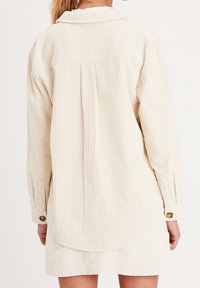 Snövit corduroy skjorta med långa ärmar, knäppning vid manschetterna och spetsig krage. Har en vertikal ribbad textur och rundad fåll.