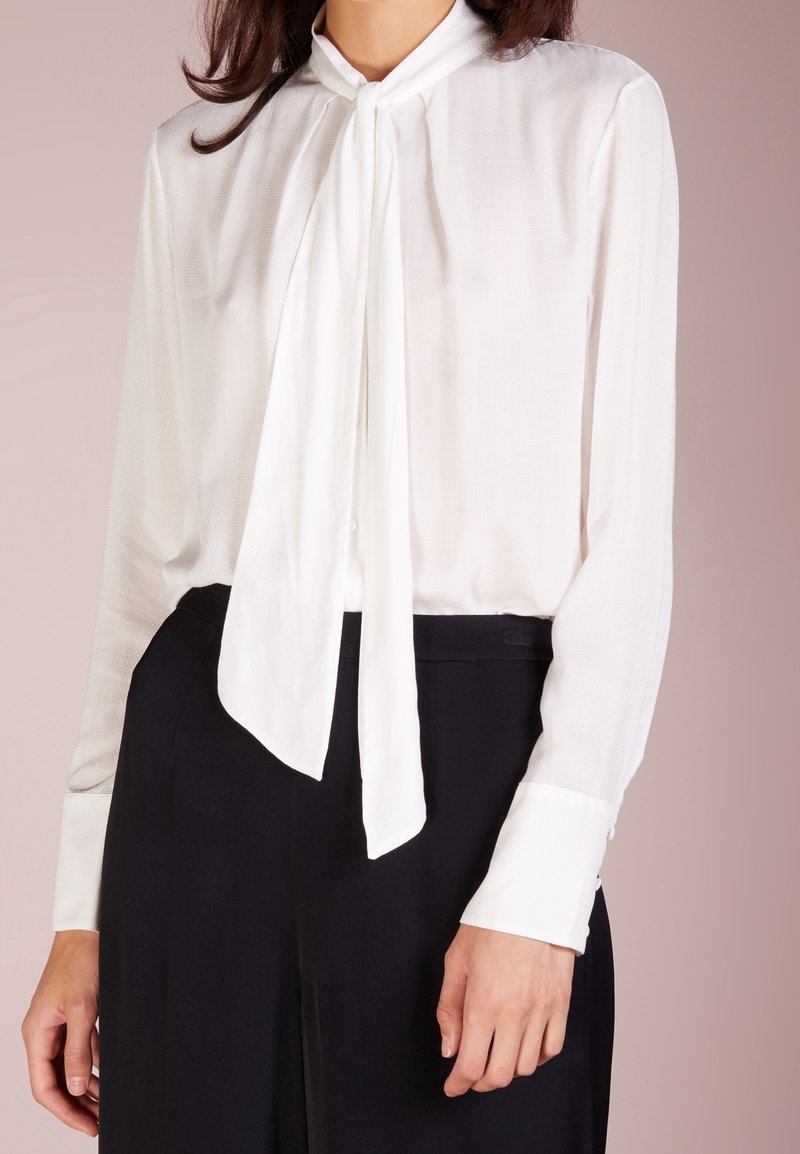 Blouse blanche à manches longues avec un détail noué au col, tissu texturé, coupe slim et poignets boutonnés. Associée à un pantalon noir taille haute.