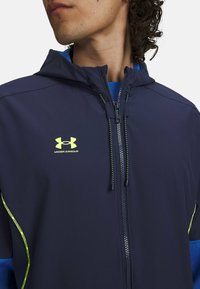 Navy hooded jacket with a zipper and lime green accents. Features Under Armour logo on chest, lightweight material, and contrasting blue inner layer.