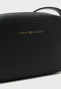 Bolso de cuero negro con superficie texturizada y logo dorado "TOMMY HILFIGER". Presenta una forma rectangular elegante y una correa delgada.