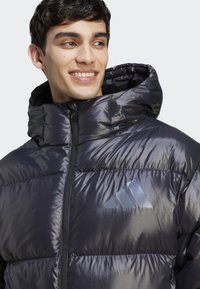 Puffetjakke i mørkegrå med skinnende, quilted tekstur. Har en lynlåslukning foran, hætte og et præget logo på brystet.