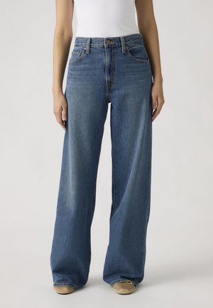 Person iført høj taljede, løstsiddende blå jeans og beige sko, stående mod en ensartet lys baggrund.