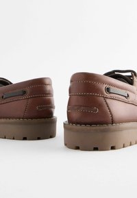 Braune Lederschuhe mit einer strukturierten Oberfläche, ausgestattet mit einer dicken Gummisohle und dekorativen Nähten. Die Rückansicht zeigt ein rundes Design.
