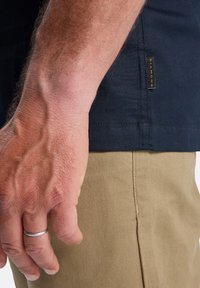 Marineblauw overhemd met korte mouwen en een gestructureerde afwerking, gecombineerd met lichtbruine katoenen broek. Een klein zwart label is zichtbaar aan de zijkant van het overhemd.