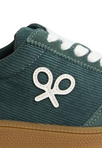 Zapatillas de pana verde oscuro con cordones blancos y un logo blanco. Presenta una parte superior texturizada y una suela de goma marrón con un patrón en su superficie.