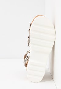 Sandale à semelle blanche avec une semelle épaisse et des brides imprimées serpent, appuyée contre un mur d'angle blanc sur un sol blanc.