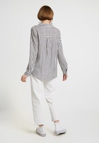 Camicia a maniche lunghe con righe verticali nere e bianche, colletto con bottoni e piega posteriore, abbinata a pantaloni bianchi corti e sneaker.
