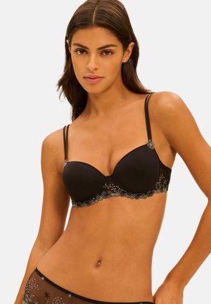 Donna che indossa un reggiseno nero e mutandine in tulle trasparente abbinate con ricami floreali, posando davanti a uno sfondo chiaro e uniforme.