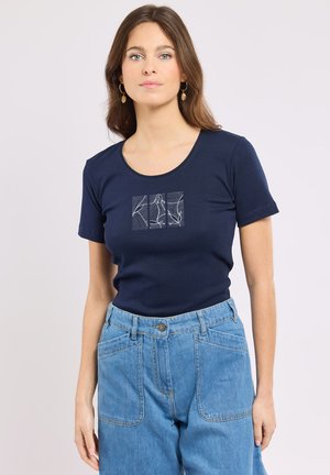 Femme portant une chemise bleu marine à manches courtes avec un motif abstrait blanc en lignes et un jean taille haute bleu clair, debout devant un fond clair.
