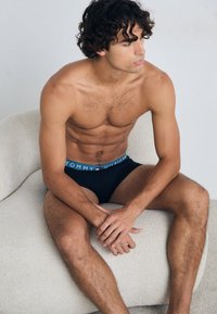 Sous-vêtements noirs avec une ceinture de marque bleue, rouge et blanche, portés par une personne assise sur une chaise texturée de couleur claire.