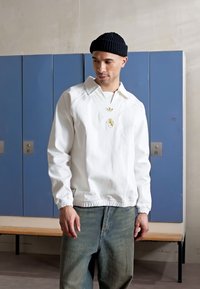 Bílá pletená bunda s límcem, elastickým pasem a dekoračním knoflíkem. Nosit s modrými džínami. Na hlavě tmavý pletený klobouk.