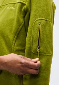 Άτομο που τραβάει το φερμουάρ στην τσέπη του μπουφάν φλις ελιάς στον άνω βραχίονα, δείχνοντας την υφή και τις λεπτομέρειες του εξωτερικού ρουχισμού.