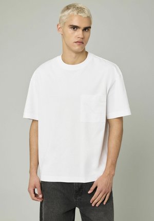T-shirt en coton blanc avec un col rond et des manches courtes. Présente une poche sur le devant et un logo de marque discret.