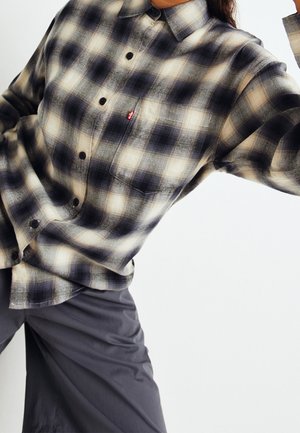 Personne portant une chemise à carreaux noir et blanc à boutons avec une poche poitrine et un pantalon sombre ample sur un fond clair uni.