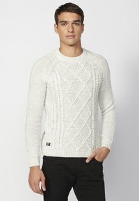 Weißer Strickpullover mit strukturiertem Rautenmuster, Rundhalsausschnitt und gerippten Bündchen. Kombiniert mit schwarzen Hosen.