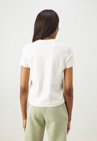 White cotton t-shirt with short sleeves and a round neckline, featuring a slightly cropped hem. Worn with light green pants.