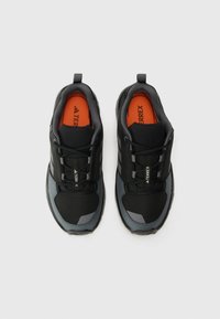 Schwarze Sportschuhe mit grauen Akzenten, strukturiertem Stoff und einer ausgeprägten Sohle. Das Innere verfügt über eine leuchtend orange Innensohle mit Branding.