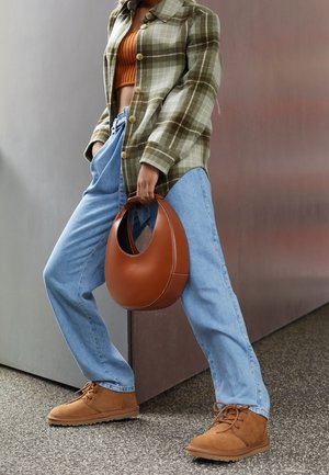 Personne portant une veste à carreaux verte, un top court orange, un jean bleu clair, des bottes en suède beige, tenant un sac à main rond en cuir marron.
