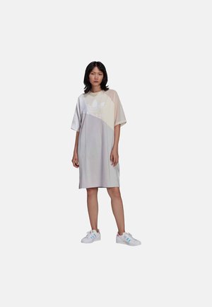 Abito oversize a maniche corte di colore grigio chiaro, con un pannello diagonale a contrasto che presenta il logo Adidas. Indossato con sneakers bianche.