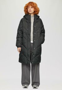 Gequiltede zwarte pufferjas met capuchon, twee voorkruiken, verstelbare trekkoord in de taille en ribbels bij de mouwen, gedragen over een grijze trui en broek.