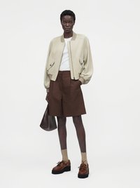 Femme portant une veste bomber beige, un haut blanc, un short marron au genou, des chaussettes beige, des chaussures compensées marron, tenant un sac foncé avec une main dans la poche.