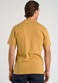 Żółta koszulka polo z krótkim rękawem o fakturowanym wzorze. Posiada klasyczny kołnierzyk i prosty dół. Materiał wydaje się gładki i lekki.
