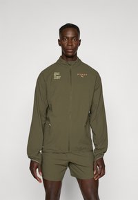 Olivengrønn treningsjakke med glidelås, høy krage og logodetaljer. Glatt tekstur, lange ermer og tettsittende design kombinert med shorts.