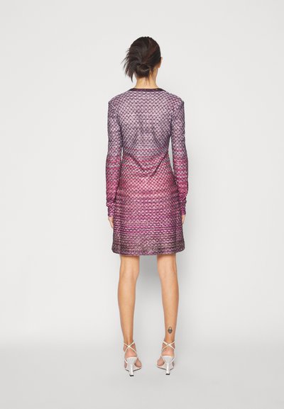 Γυναίκα με χτένισμα σε σινιόν, φοράει ένα μακρυμάνικο, υφασματοποιημένο μωβ-ροζ φόρεμα και ασημί τακούνια, στέκεται μπροστά σε έναν απλό ανοιχτό φόντο.