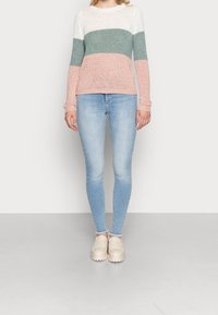 Gestreifter Strickpullover mit weißen, grünen und pinken horizontalen Streifen, kombiniert mit hellblauen Skinny-Jeans und beigen Slipschuhen.