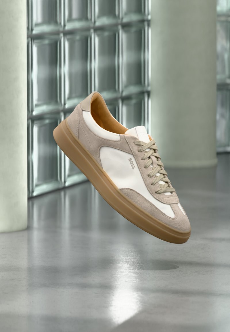 Baskets beige et blanches avec semelle marron clair et logo « BOSS », suspendues au-dessus d’un sol lisse et réfléchissant avec un mur en parpaings de verre en arrière-plan.