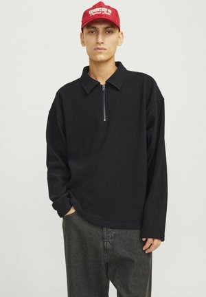 Black ribbed long-sleeve pullover with a zipper collar and relaxed fit, paired with dark gray pants. Accent of red cap with text.