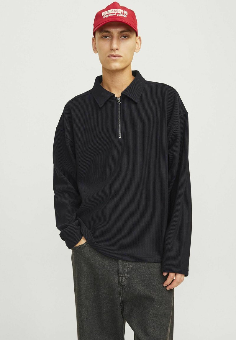 Pull noir à manches longues et côtes, avec un col zippé et une coupe décontractée, associé à un pantalon gris foncé. Accent d'une casquette rouge avec du texte.