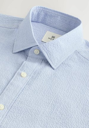 Chemise texturée bleu clair avec un col boutonné, dotée de deux boutons blancs et d'une étiquette avec le nom de la marque à l'intérieur du col.